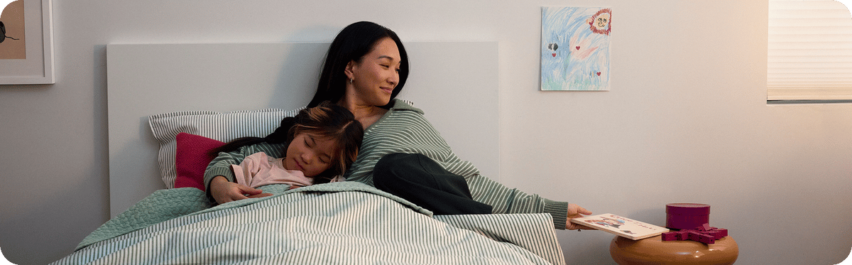 Woman and child lying in bed together, reading a book in a cozy bedroom setting.