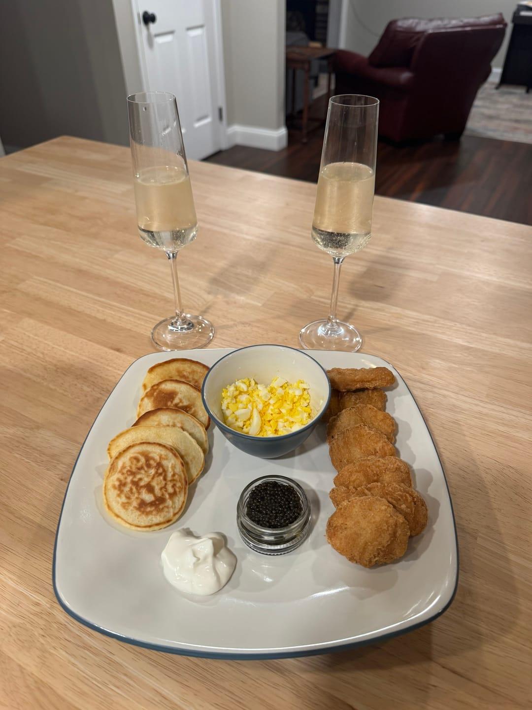 Customer's Siberian Sturgeon Caviar served with pancakes, eggs, and sides on a white plate with two champagne glasses.
