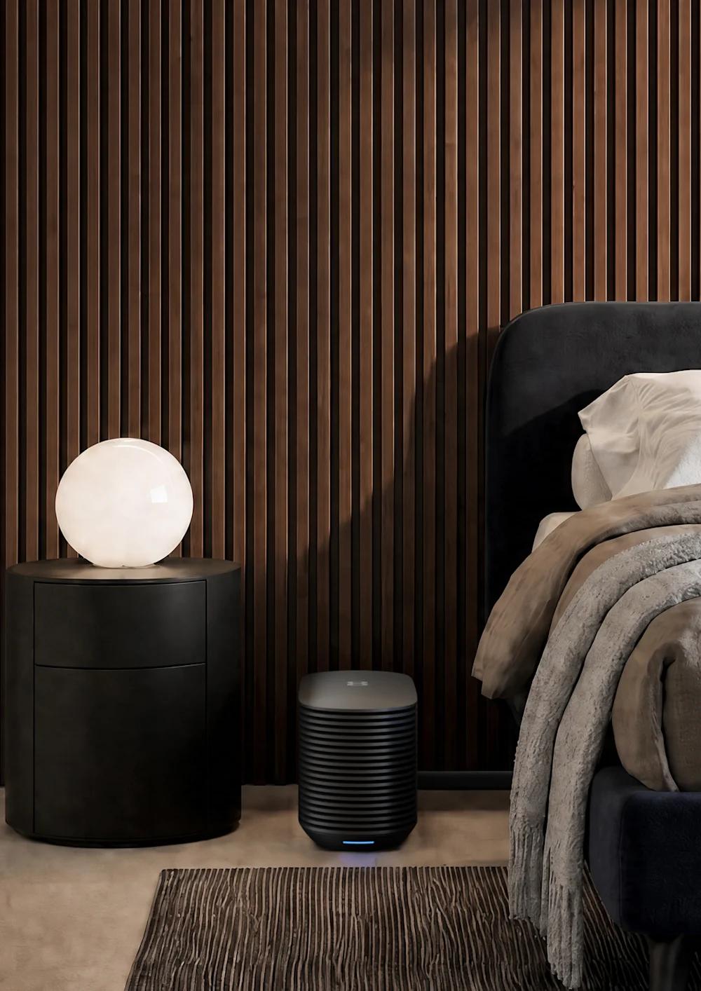 A couple in a sunlit bedroom, one sitting on the edge of the bed and the other reclining in a lounge chair, with the Pod Hub on the floor beside the nightstand