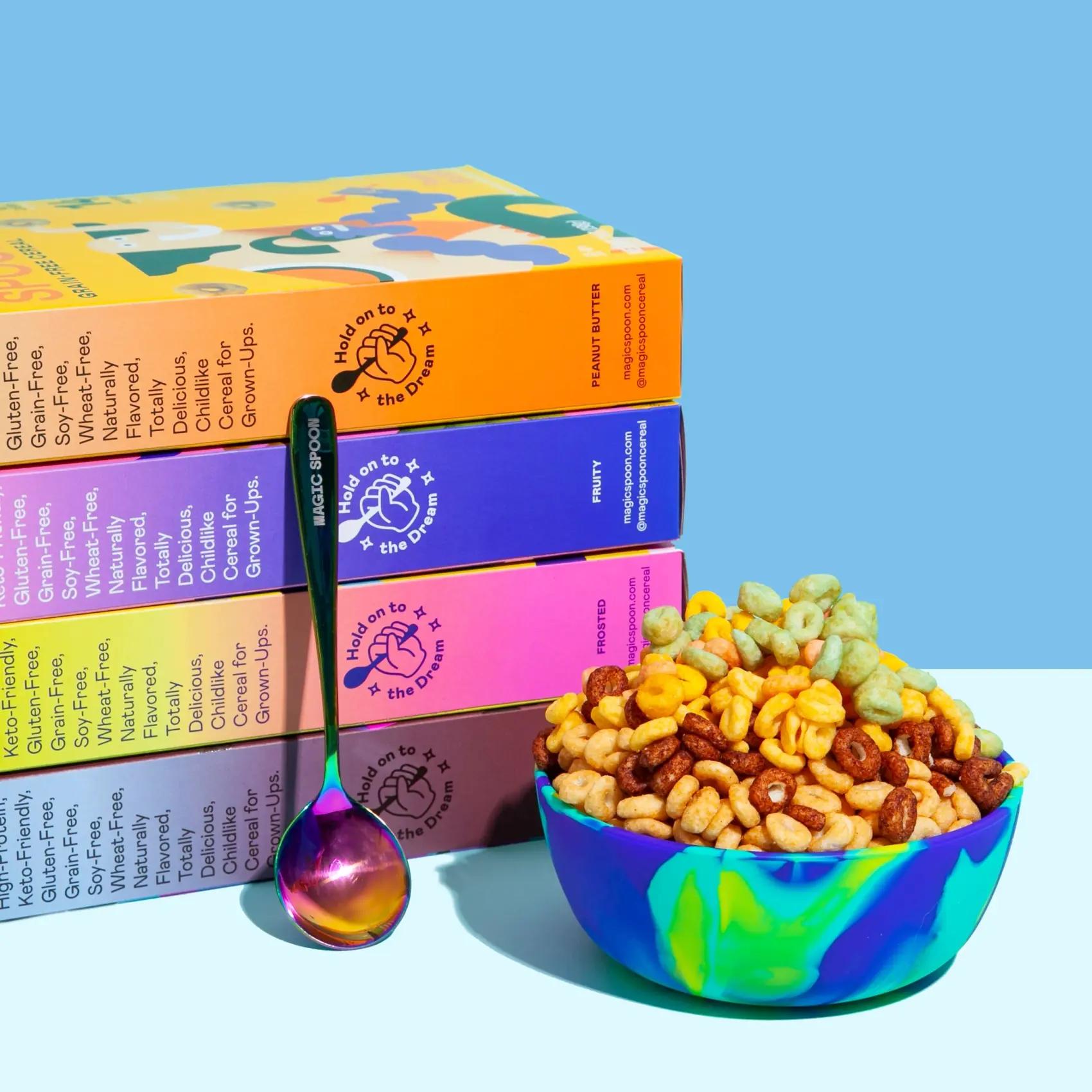 Stack of Magic Spoon cereal boxes in Peanut Butter, Fruity and Frosted beside an iridescent spoon and a swirled bowl of mixed cereal