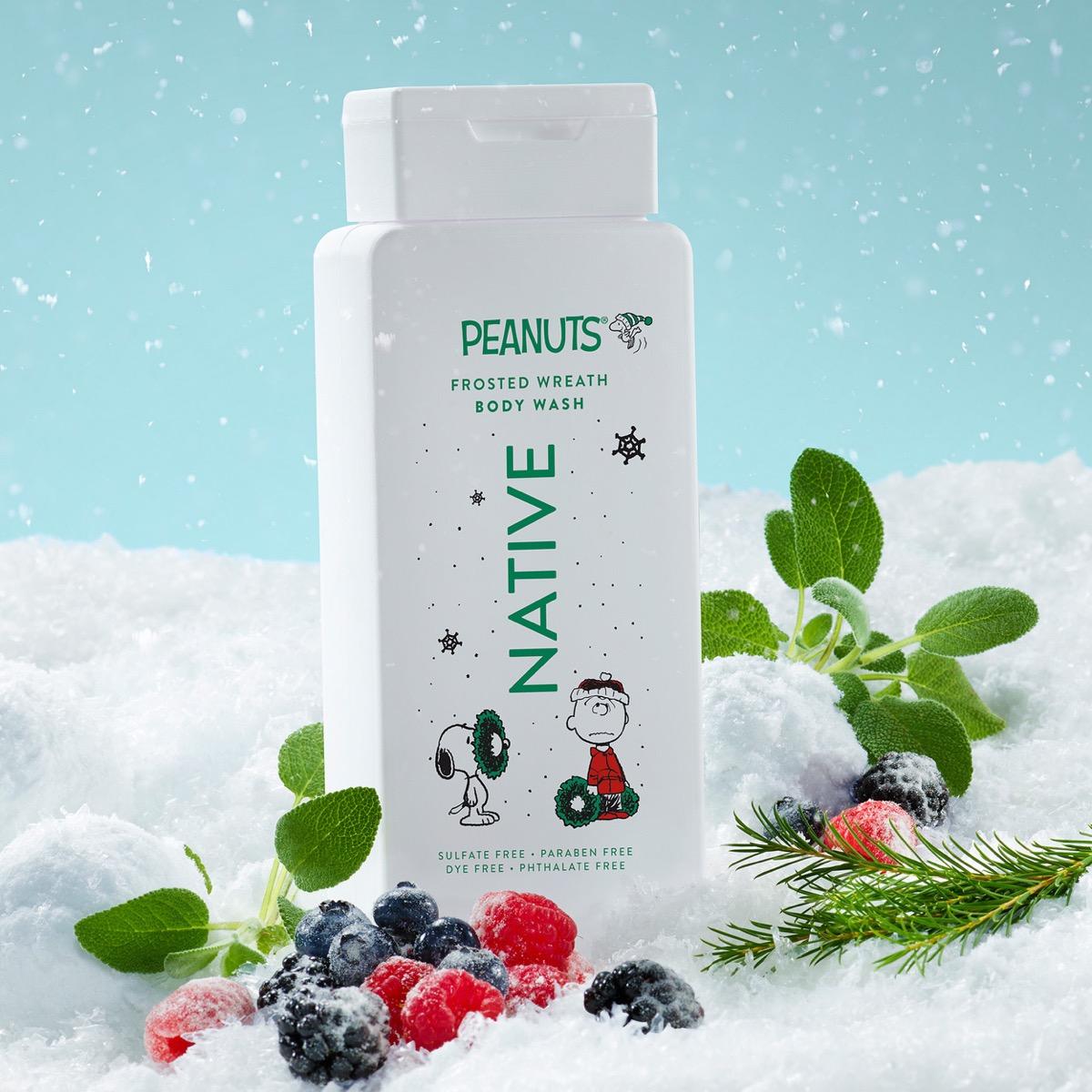 A bottle of Peanuts 'Frosted Wonderland' body wash stands in a snowy scene with berries, green leaves, and pine branches. The bottle is white with green and black text and cartoon graphics of Snoopy and Charlie Brown.