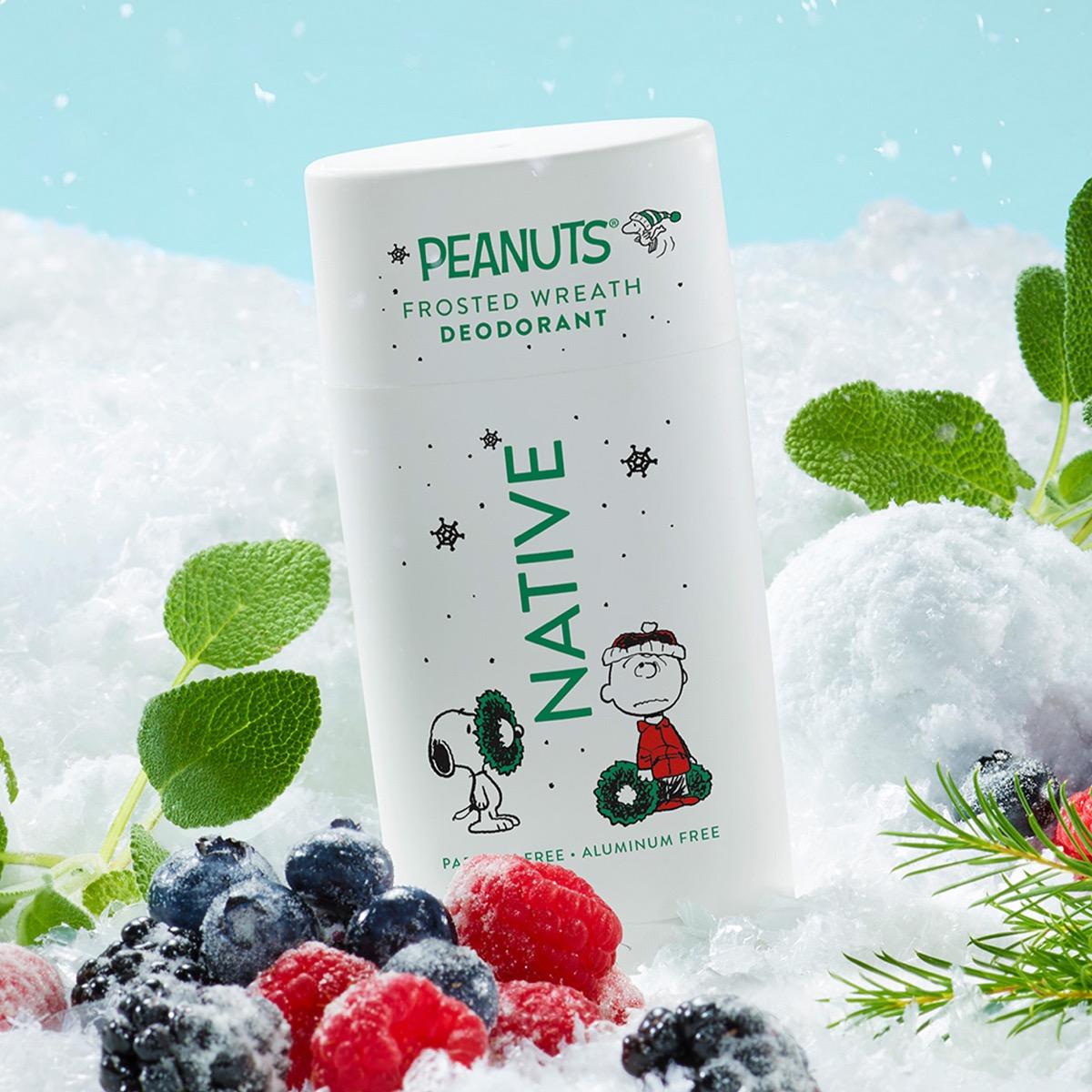 A white deodorant stick labeled PEANUTS FROSTED WREATH DEODORANT with Snoopy and Woodstock graphics, standing in snow with berries and mint leaves around it.