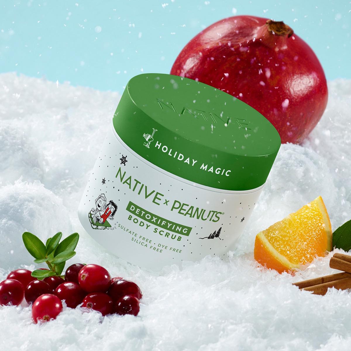 A tub of Native x Peanutz Holiday Magic Detoxifying Body Scrub sits in faux snow, styled with a pomegranate, orange slice, cranberries, and cinnamon sticks against a light blue background.