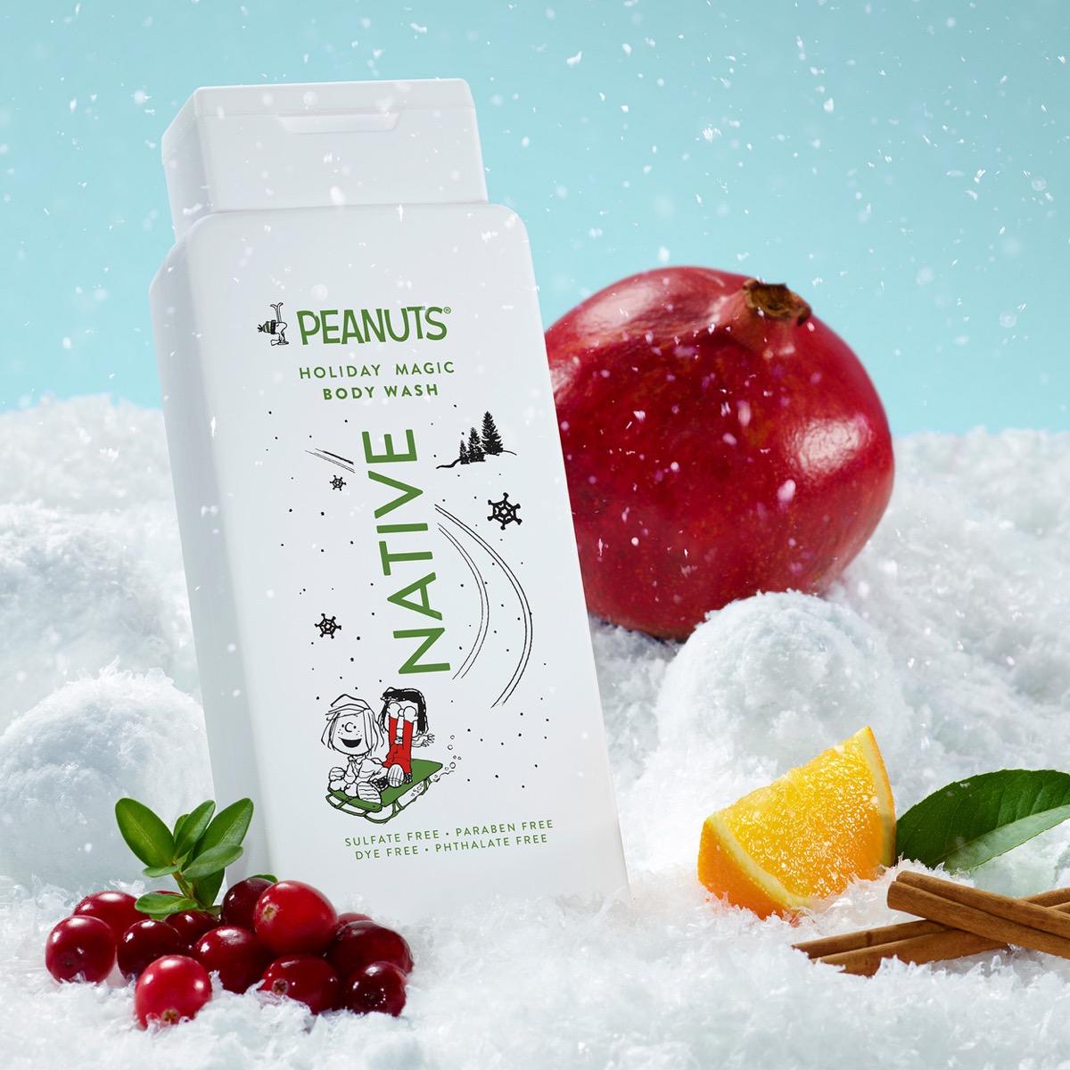 A bottle of PEANUTS Native Holiday Magic Body Wash stands in white faux snow with a red pomegranate, cranberries, orange slice, cinnamon sticks, and green leaves around it against a light blue background.