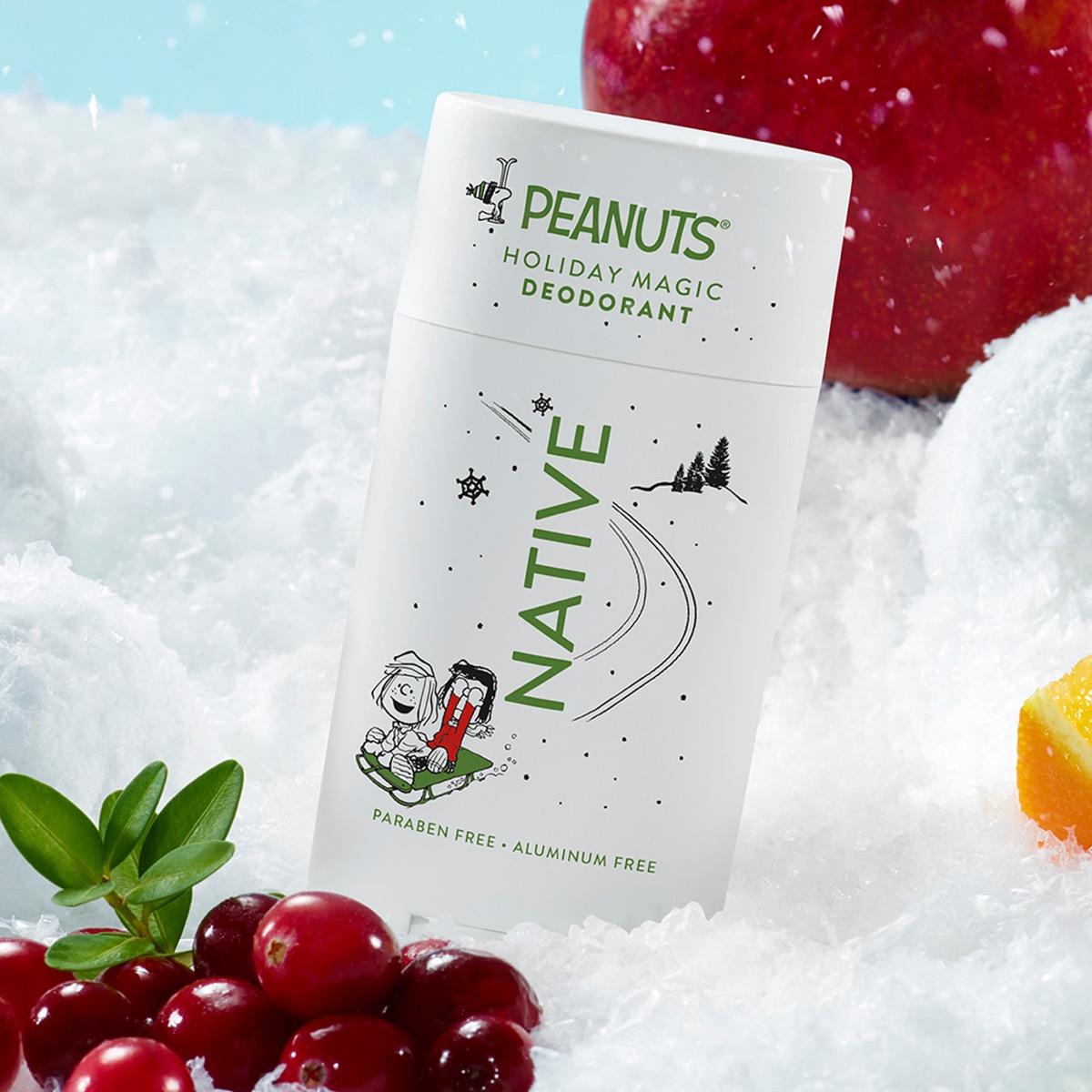 A white Peanuts Holiday Magic Deodorant stick labeled Native, surrounded by red cranberries, green leaves, a red apple, and orange slices on a bed of white snow-like texture.