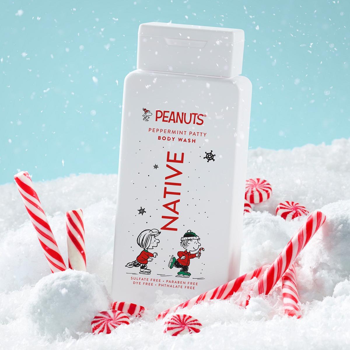 A white bottle of Native Peanuts Peppermint Patty Body Wash stands upright in a snowy scene with red-and-white candy canes. The bottle features the Peanuts logo and cartoon characters, with a light blue sky background and falling snow.