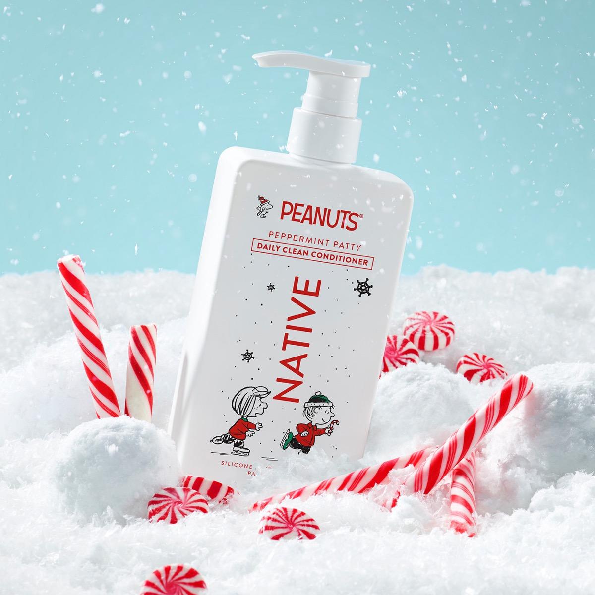 A bottle of Peanuts Peppermint Patty Daily Clean Conditioner stands in a snowy scene with candy canes. The bottle is white with red and black text and illustrations, set against a light blue sky background with falling snow.
