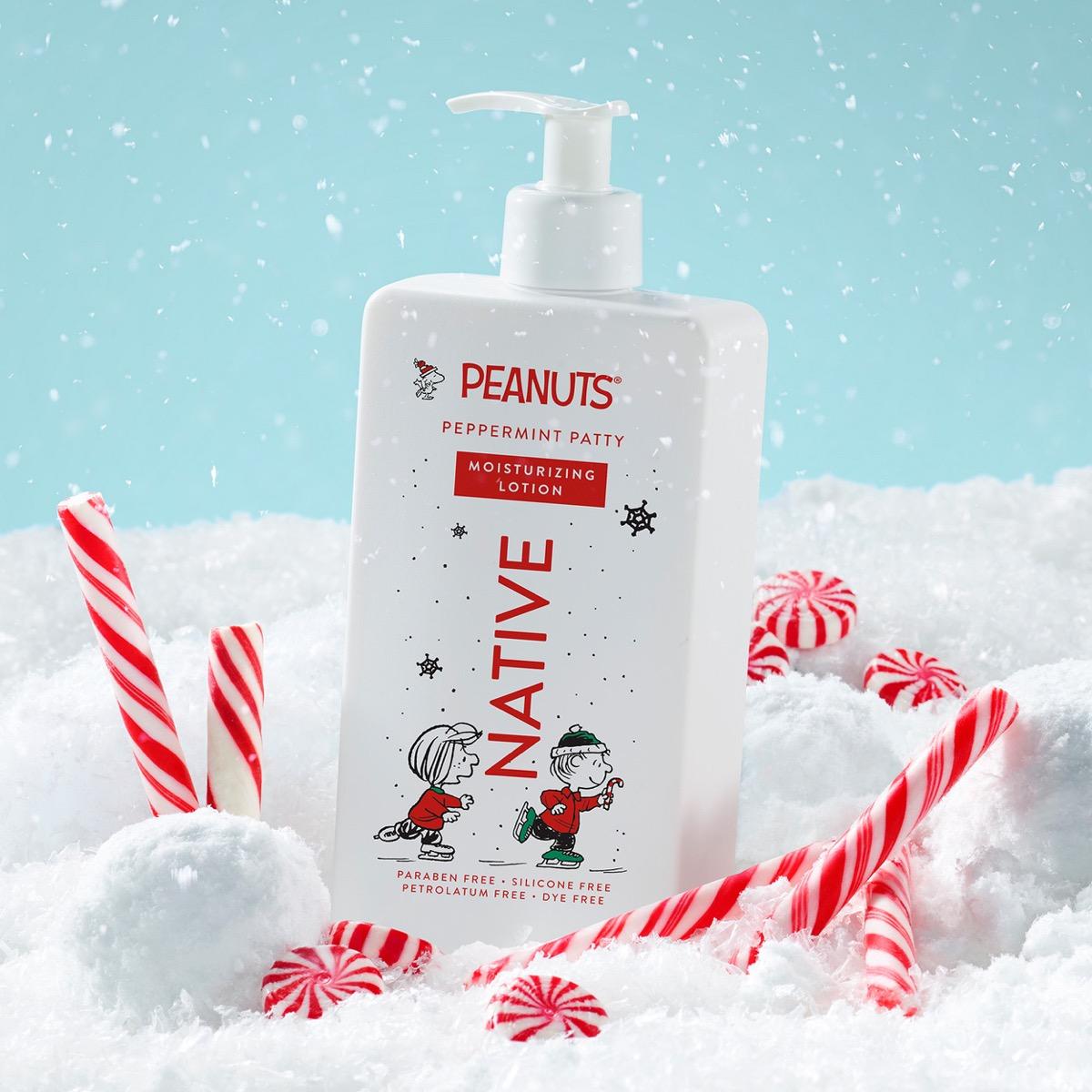 A bottle of Peanuts Native Peppermint Patty moisturizing lotion stands in a snowy scene with red-and-white candy canes. The bottle is white with holiday-themed illustrations and text, and the background is a light blue sky with falling snow.