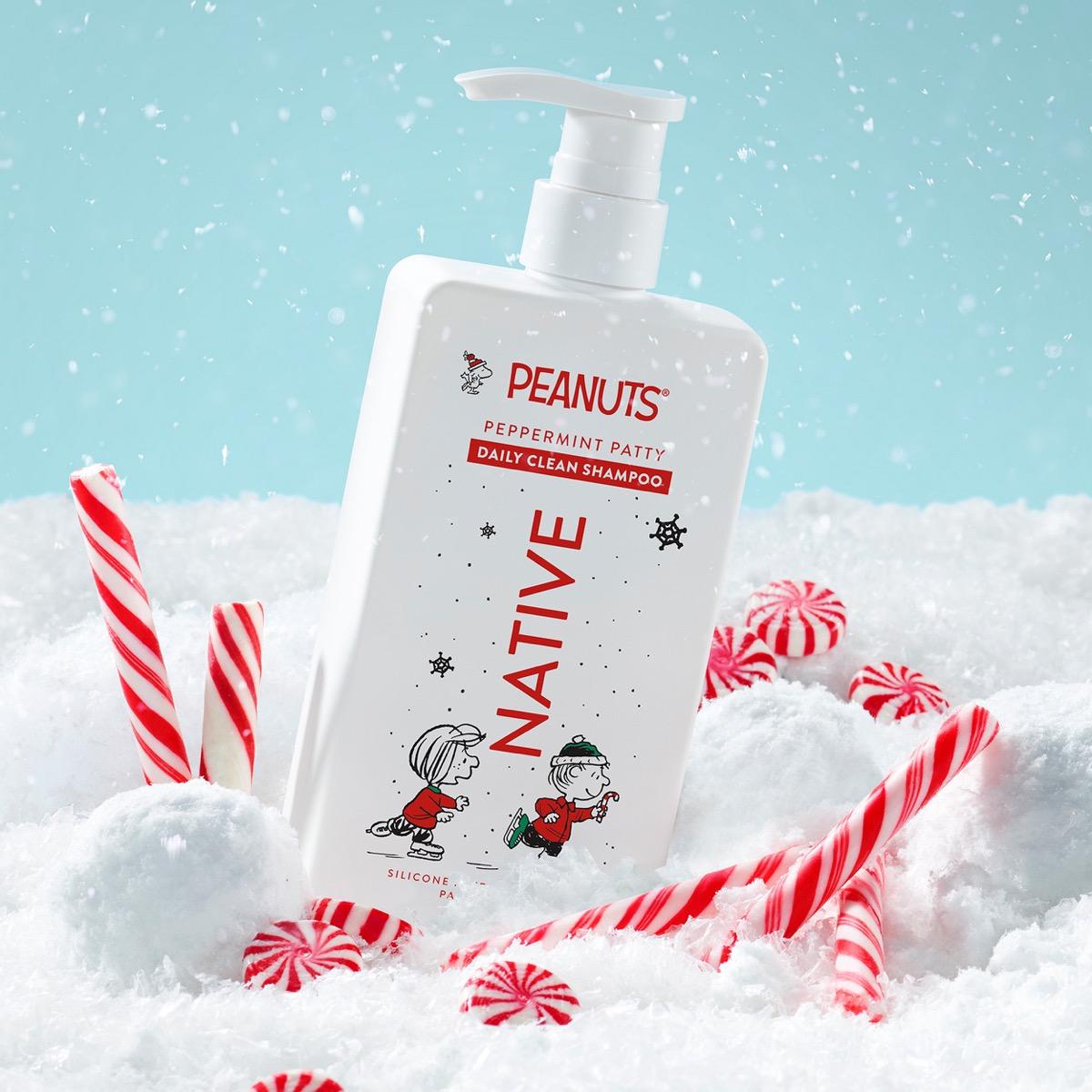 A bottle of Peanuts Peppermint Patty Daily Clean Shampoo stands in a snowy scene with red-and-white candy canes and peppermint candies. The bottle is white with red and black text and cartoon graphics, set against a light blue background with falling snow.