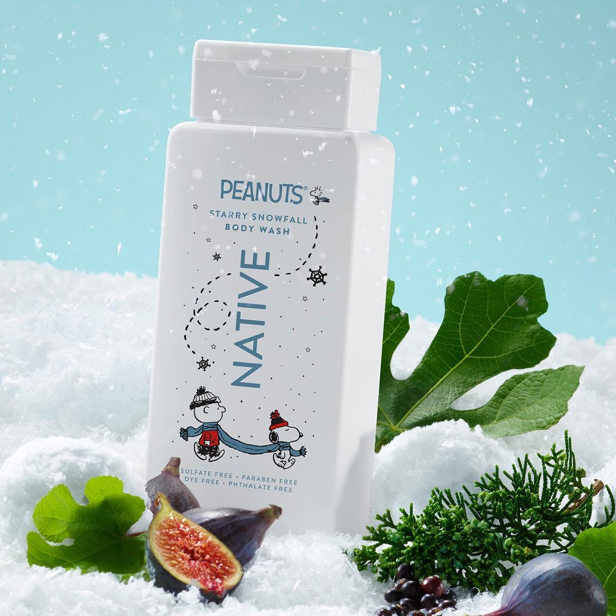 A bottle of Peanuts 'Starry Snowfall' body wash stands in a snowy scene with green leaves, figs, and pine branches. The bottle is white with blue text and Snoopy and Charlie Brown illustrations, set against a light blue background with falling snow.