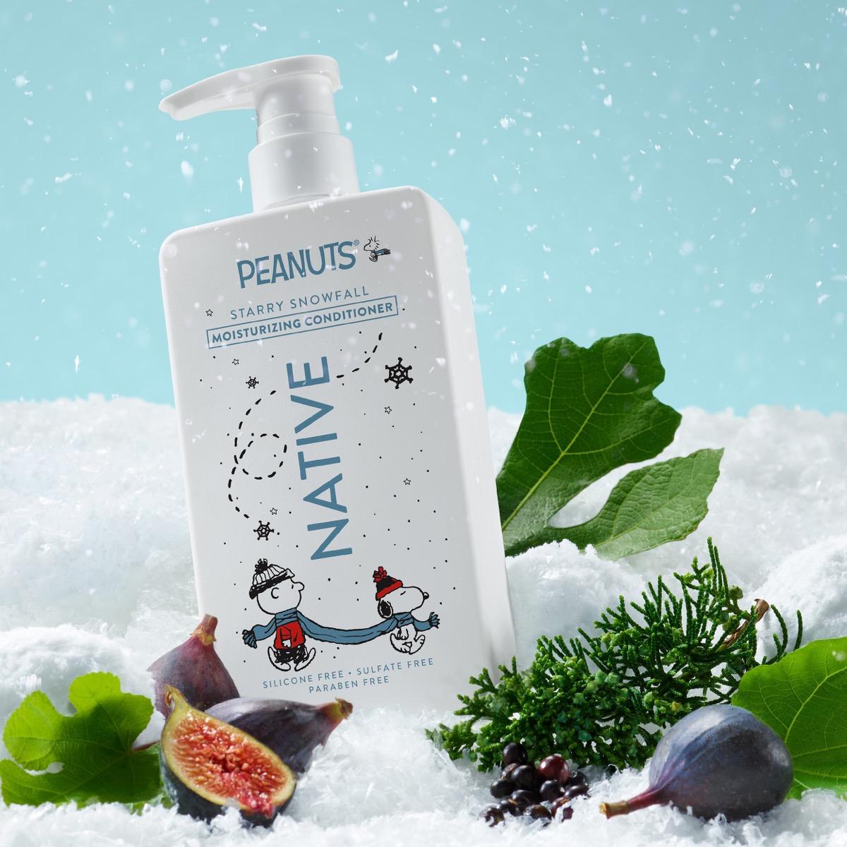 A bottle of Peanuts Native Starry Snowfall moisturizing conditioner stands in a snowy, winter-themed setup with figs, evergreen branches, and a light blue sky background.
