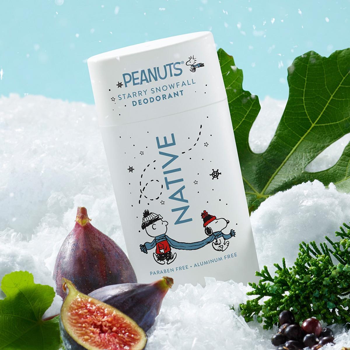 A tub of Peanuts Starry Snowfall Deodorant standing in a snowy setup with figs, green leaves, and pine sprigs. The container is white with blue and black Peanuts-themed graphics and text including 'NATIVE' and 'PEANUTS STARRY SNOWFALL DEODORANT'.