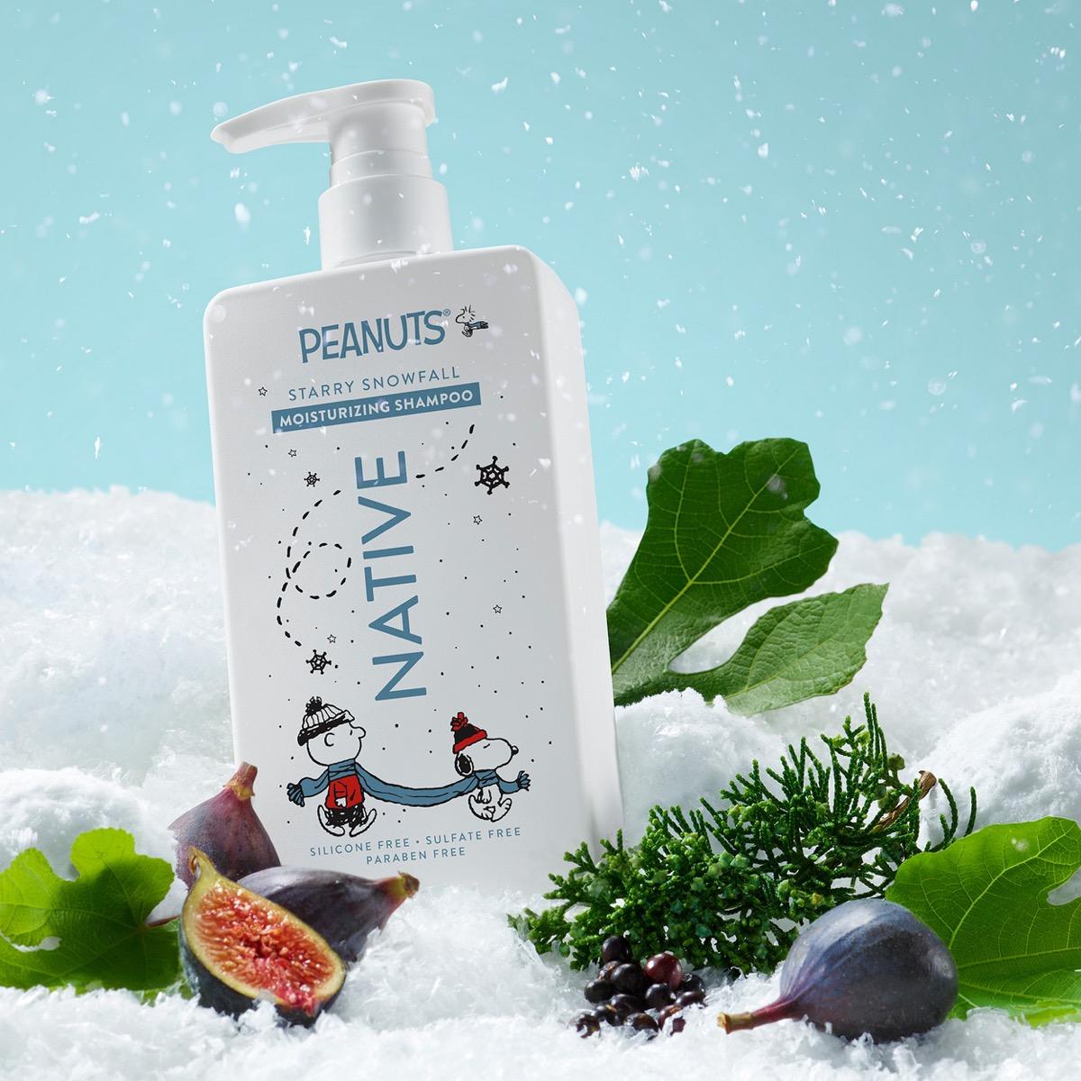 A bottle of PEANUTS Native Starry Snowfall Moisturizing Shampoo stands in a snowy scene with figs, green leaves, pine branches, and a light blue sky background. The bottle is white with blue and red graphics and text, and the label reads 'PEANUTS NATIVE STARRY SNOWFALL MOISTURIZING SHAMPOO'.