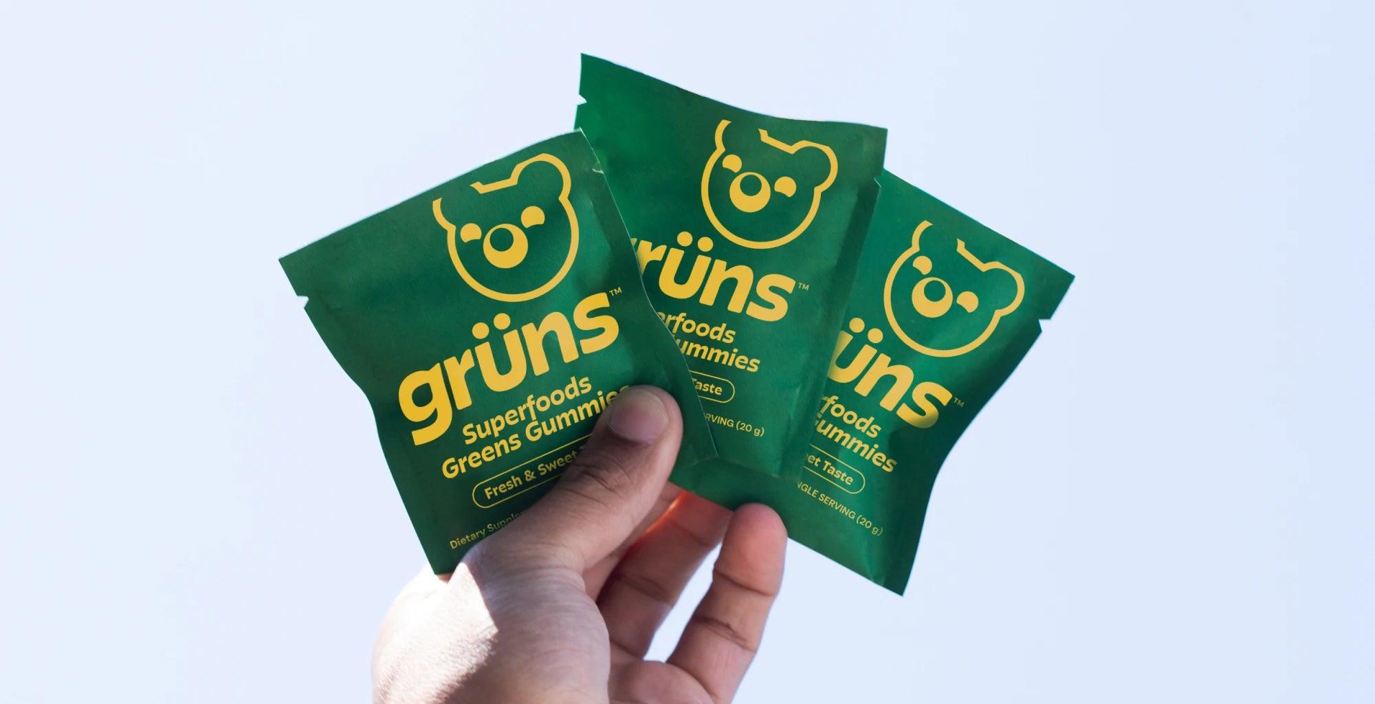 Three green packets of Grüns Superfoods Greens Gummies held by a hand against a light blue background.