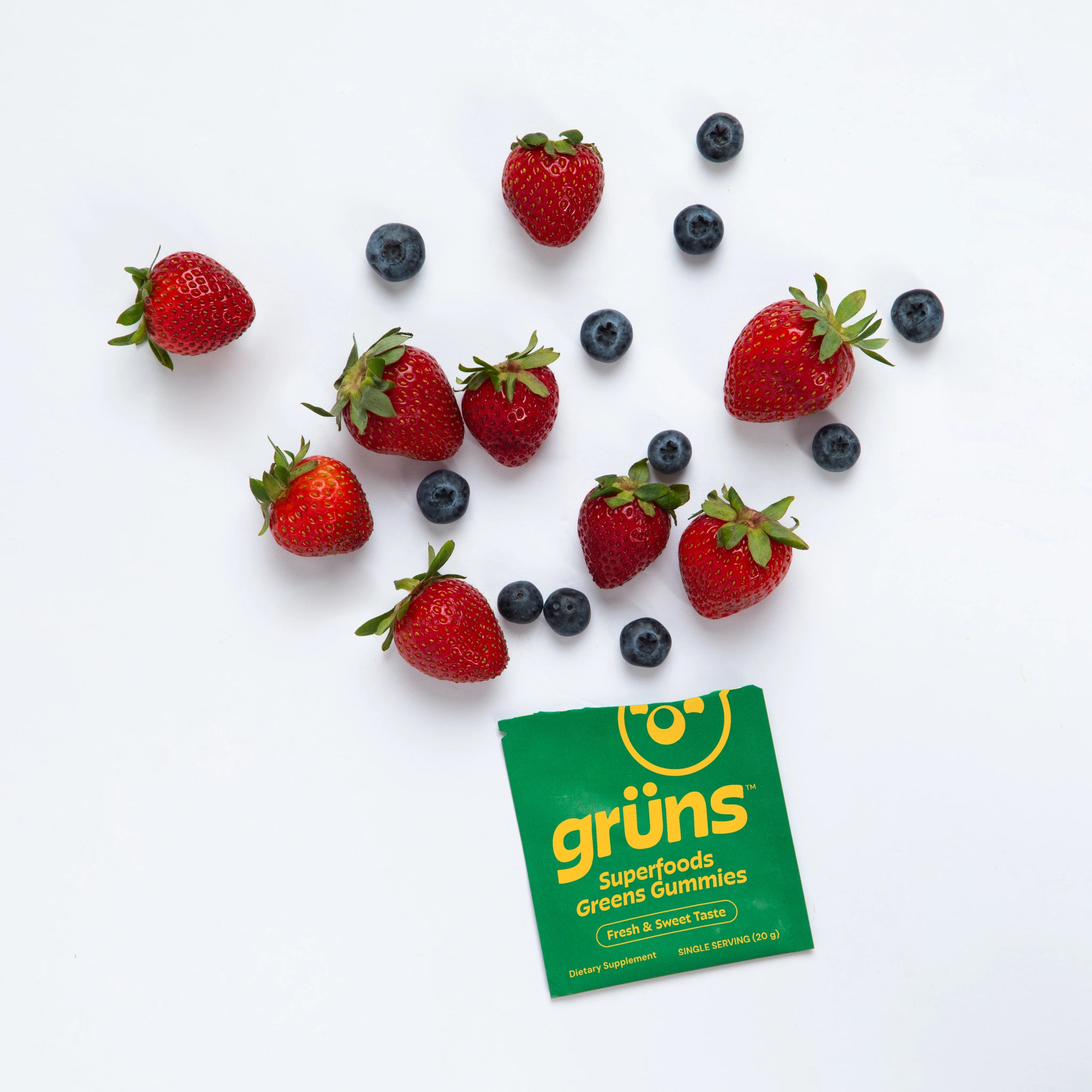 A Grüns Superfoods Greens Gummies packet surrounded by fresh strawberries and blueberries.