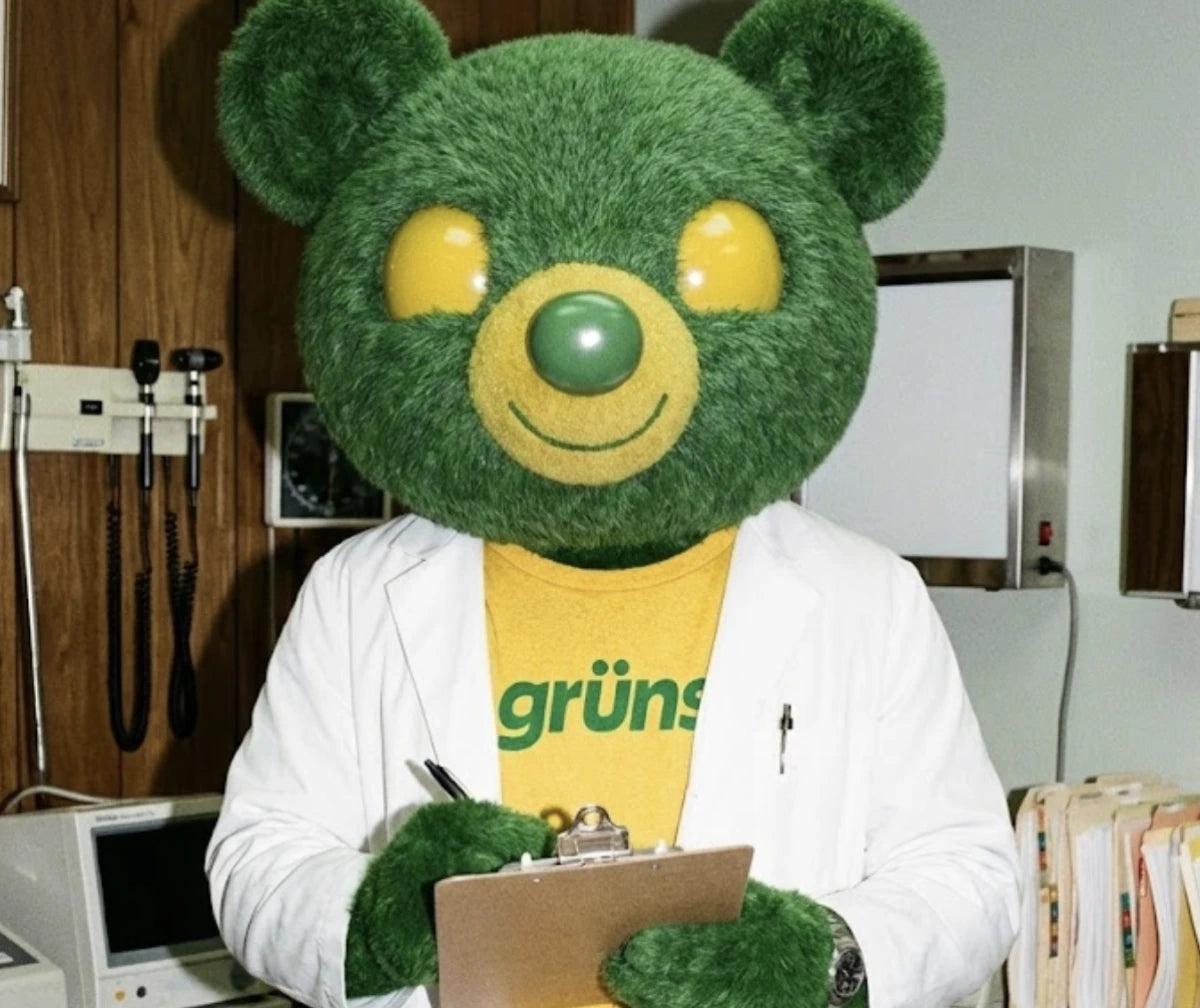 A green Grüns bear mascot in a lab coat holding a clipboard in an exam room.