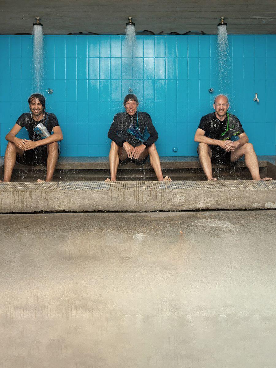 The three On Co-Founders sitting under a blue outdoor shower with water pouring down over them.