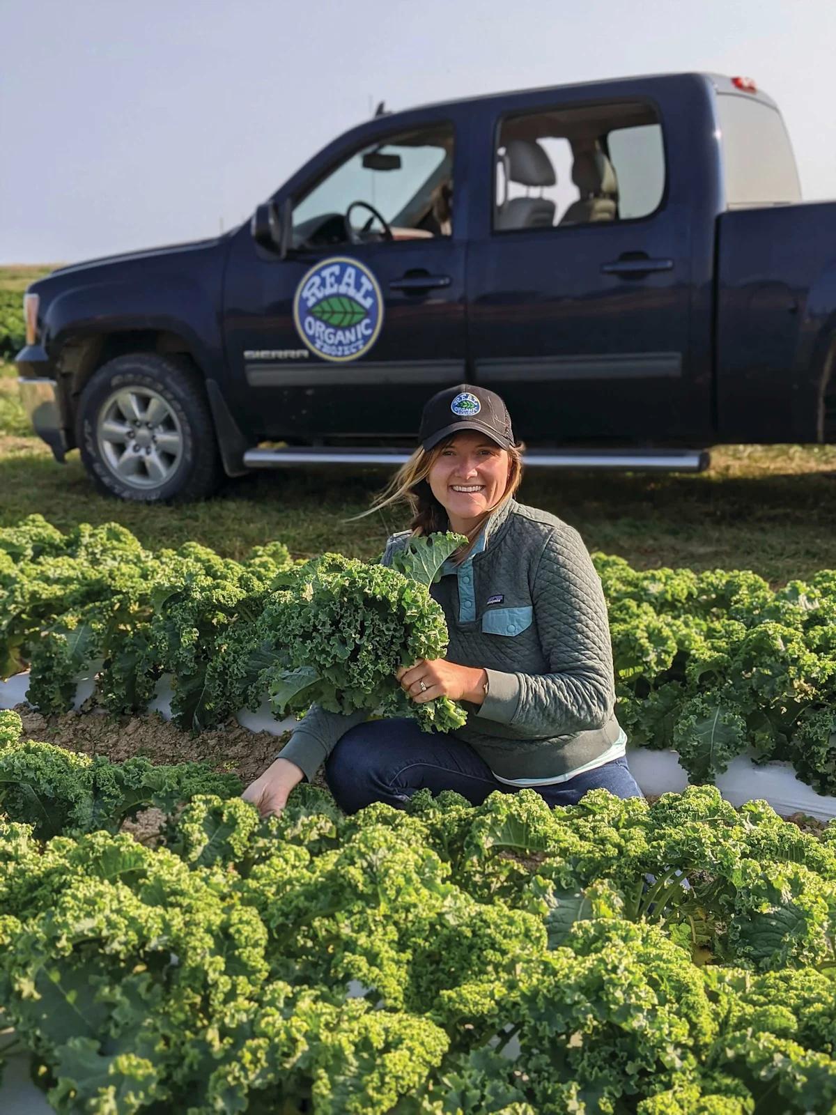 Farmer kneeling in a kale field holding a bunch of kale in front of a pickup truck at Lady Moon Farms