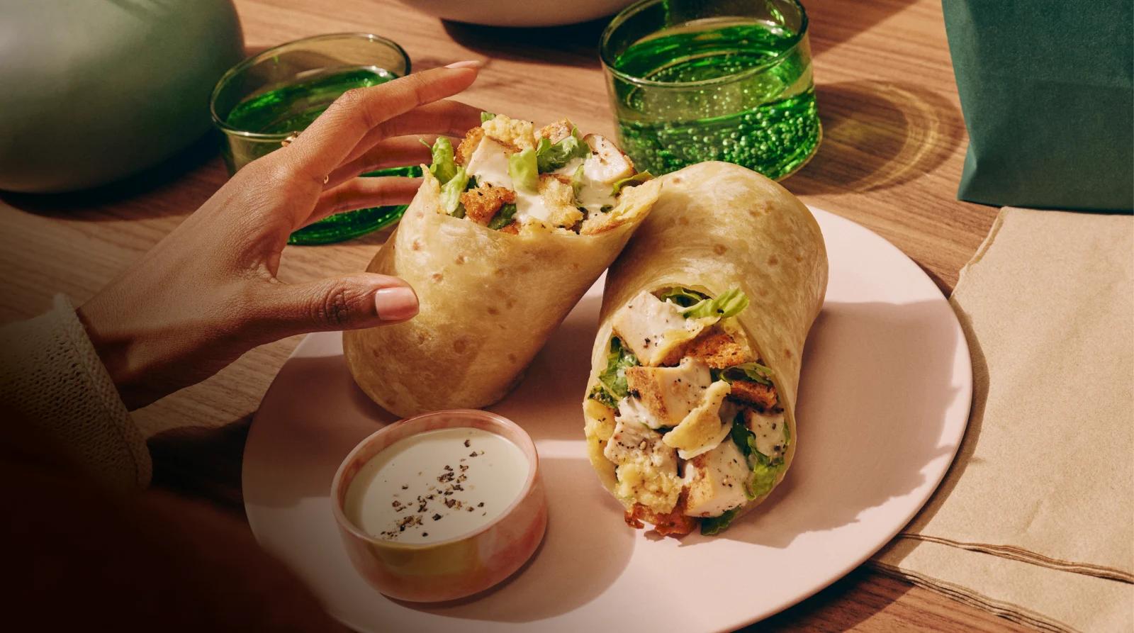 Hand picking up a Classic Chicken Caesar wrap on plate with caesar dressing on the side