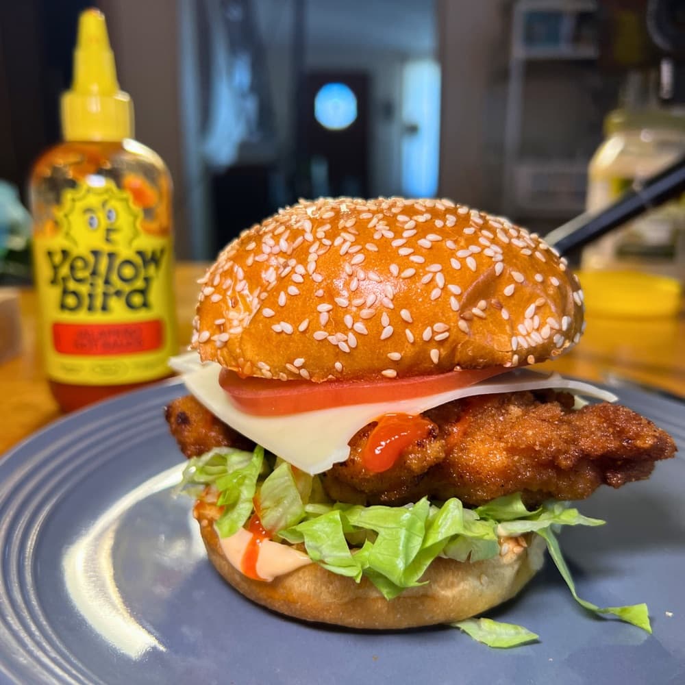 A spicy chicken sandwich on a toasted sesame brioche bun on a plate. There is a bottle of Yellowbird Jalapeño Hot Sauce in the background.