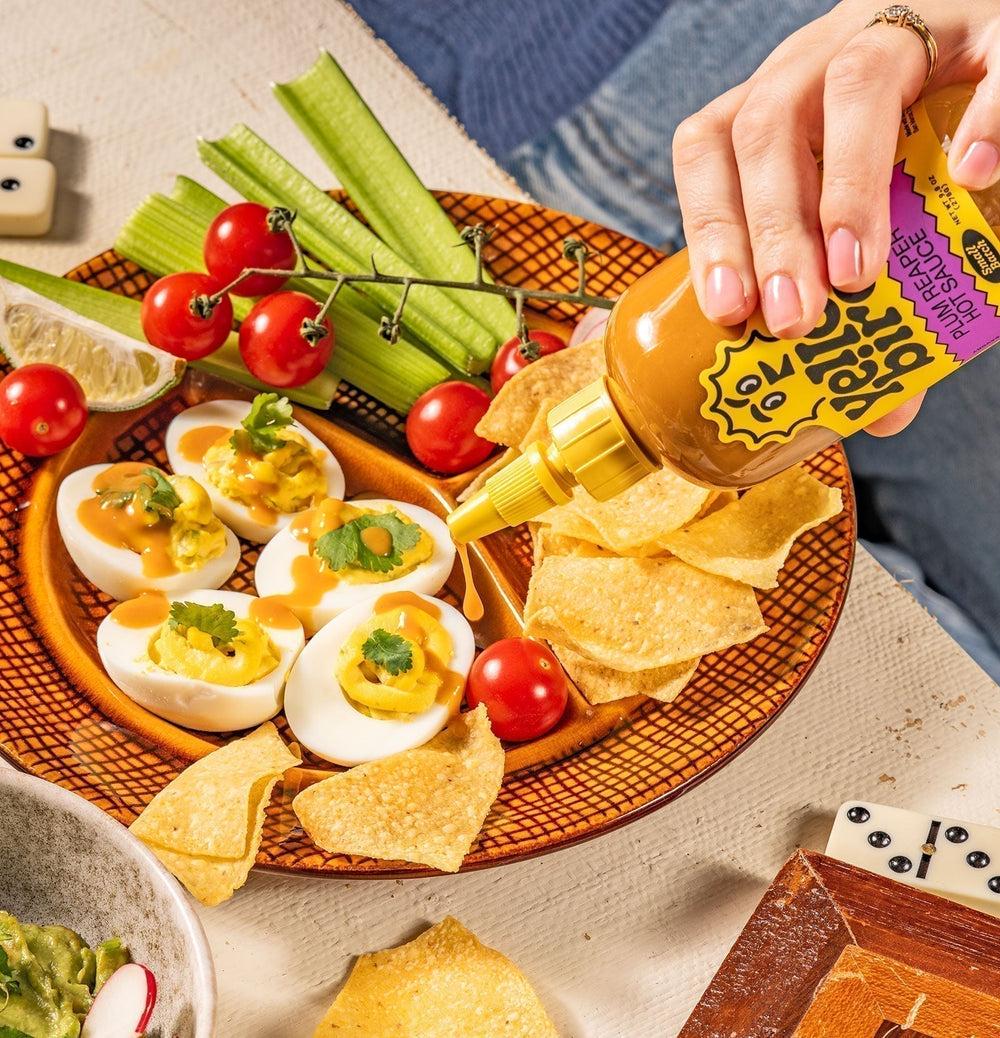 A plate containing deviled eggs, cherry tomatoes, celery, and tortilla chips. Yellowbird Plum Reaper Hot Sauce is being squeezed onto the deviled eggs.