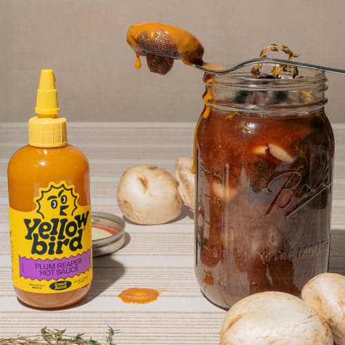 A bottle of Yellowbird Plum Reaper Hot Sauce next to a mason jar filled with spicy pickled mushrooms.
