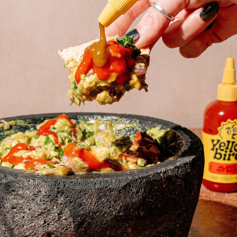Guacamole in a stone mortar, drizzled with Yellowbird Jalapeño Hot Sauce. A hand holds a chip above, loaded with guac, and drizzling Yellowbird Serrano Hot Sauce onto the chip.