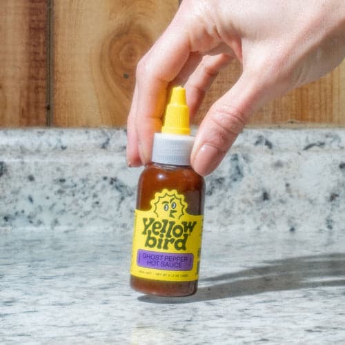 A picture of a Yellowbird Classic Ghost Pepper Hot Sauce in the 2.2 ounce size being held up by a persons fingers