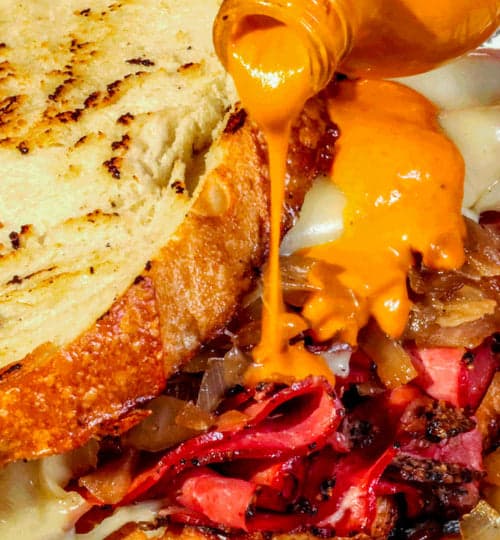 A picture of Yellowbird habanero hot sauce being drizzled on an open pastrami deli sandwich
