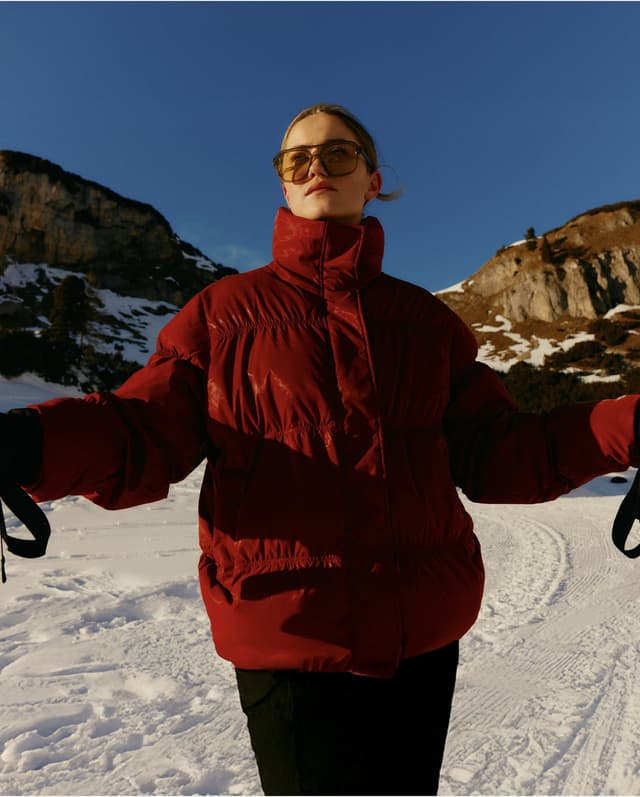 A person in a red puffer jacket stands in a snowy landscape with mountains in the background.