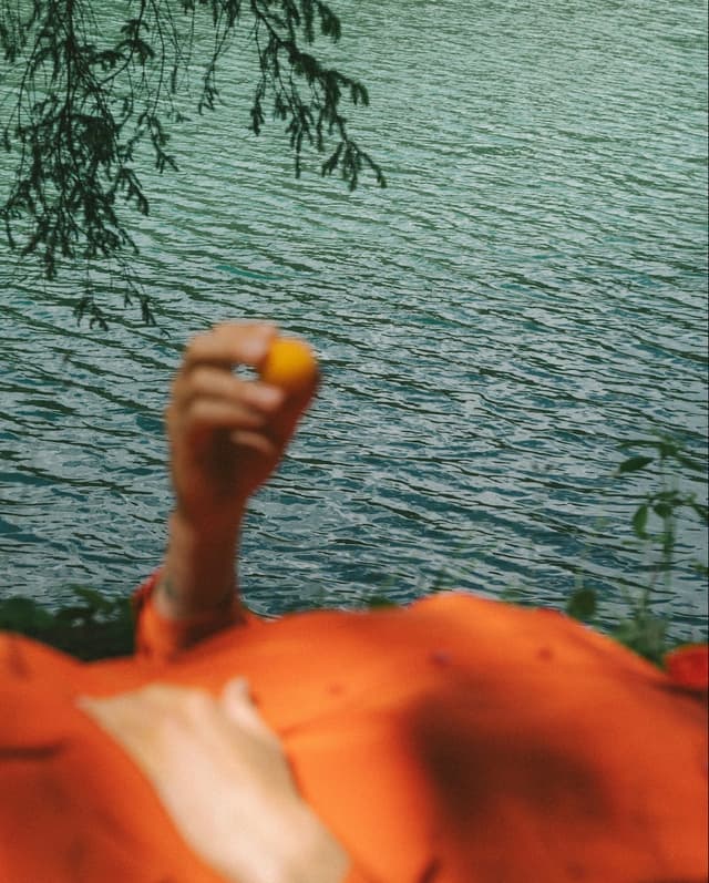 A hand holding a small yellow object above a vibrant orange garment, with water in the background.